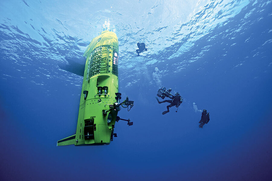A bright yellow, vertical submarine hovering in the ocean. There are scuba divers with cameras and other technology swimming around the submarine.