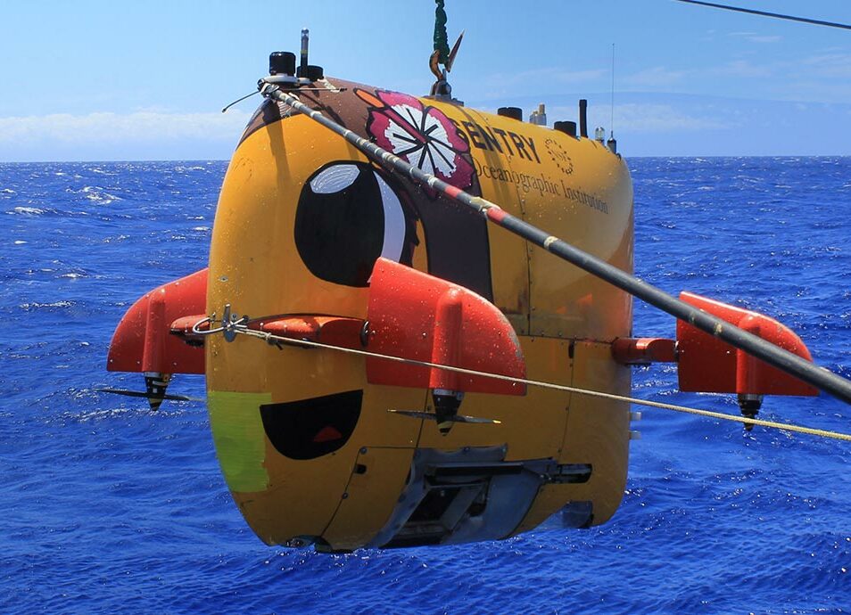 A yellow autonomous underwater vehicle being hovered overboard - soon to be submerged in the ocean.