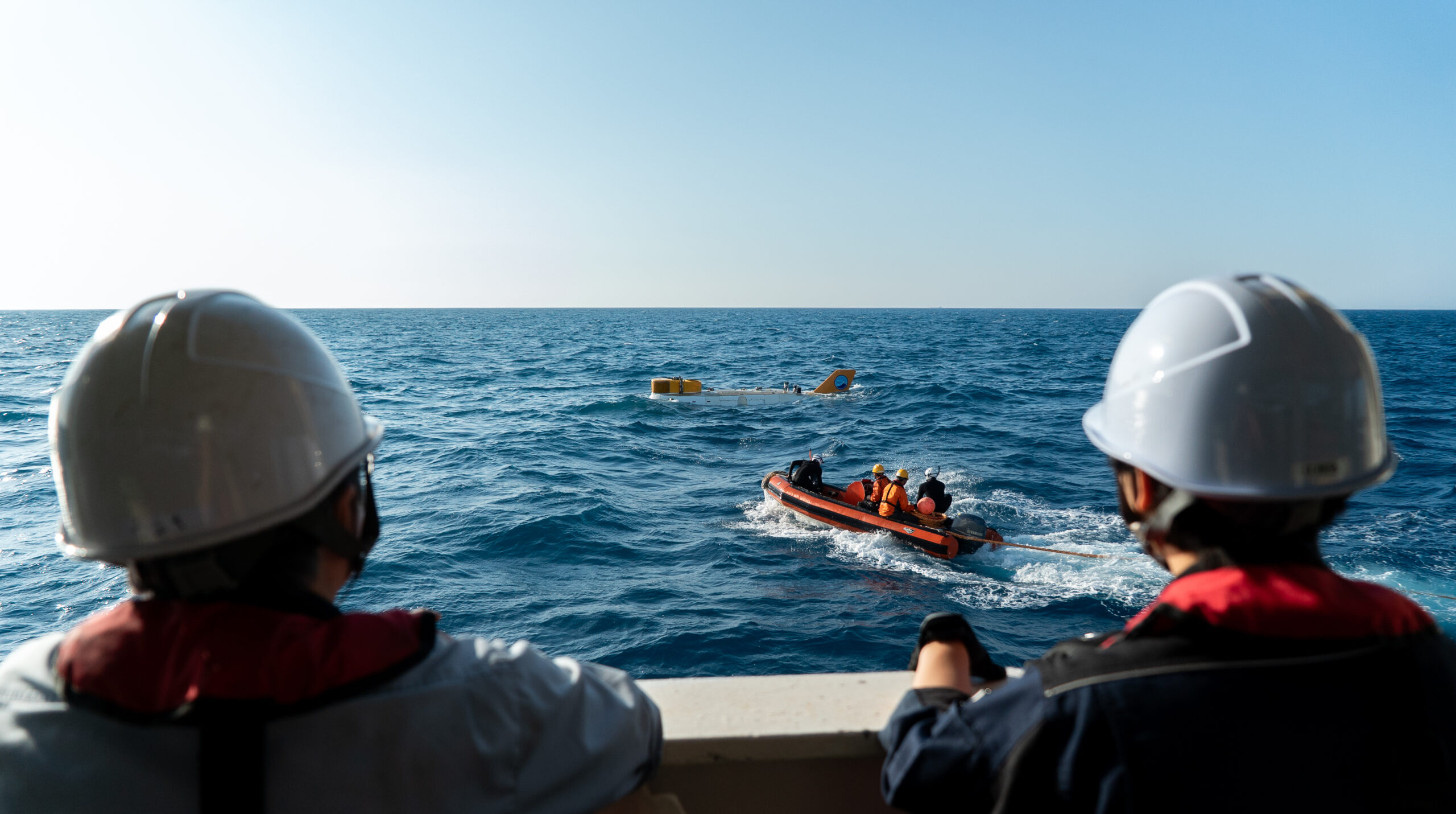 Two crew members watch from the deck as a small inflatable boat approaches an oceanographic instrument floating on the surface. The recovery is part of a broader deep-sea expedition at a dive site in the Nankai Trough, supporting ongoing marine biodiversity research.