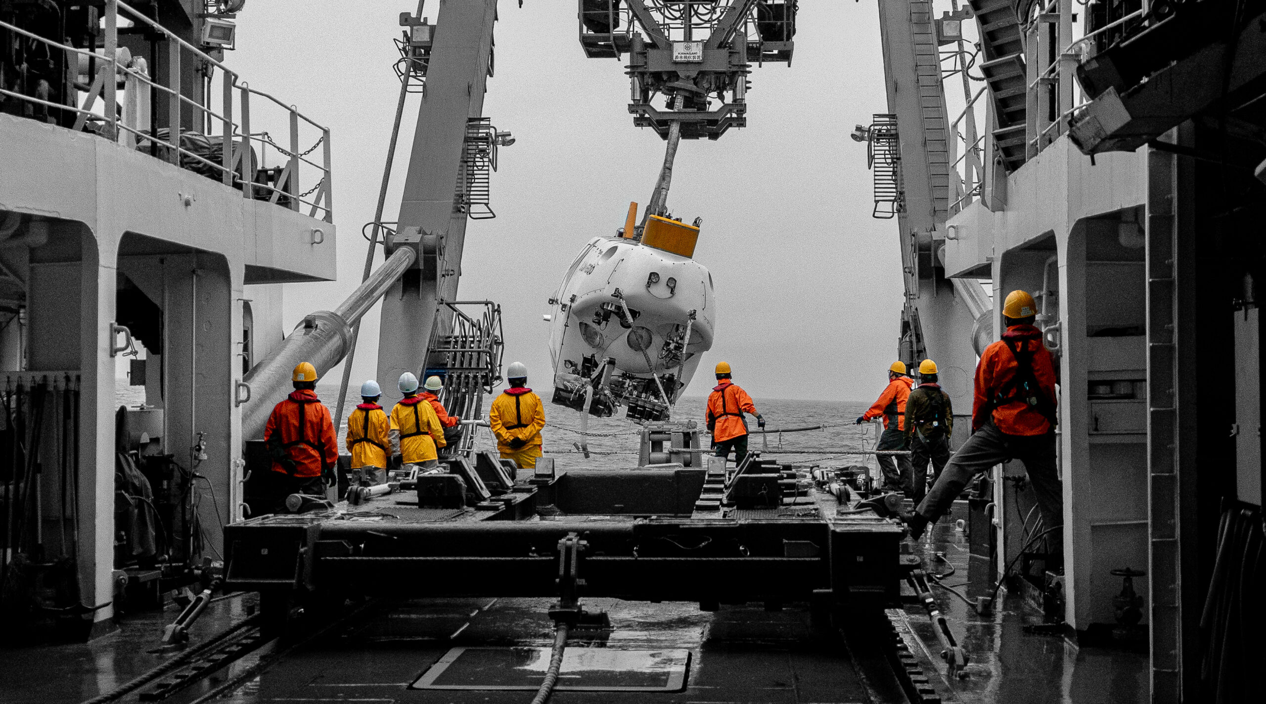 Crew members in brightly colored safety gear guide a deep-sea submersible being lowered into the ocean from a research vessel during a scientific operation. The submersible is suspended mid-air by a large deployment crane as it prepares to descend to a designated dive site in the Nankai Trough, a geologically active trench off Japan’s coast. The operation is part of a deep-sea expedition focused on exploring and documenting marine biodiversity in extreme ocean environments.